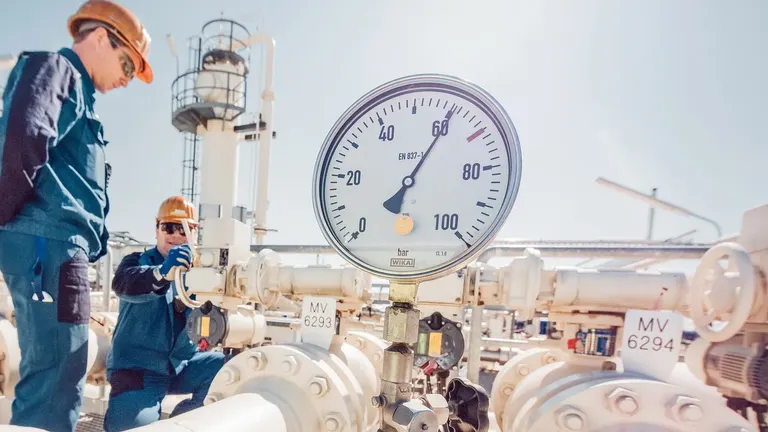Two workers in protective gear inspecting gas pipeline equipment with a large pressure gauge in focus.